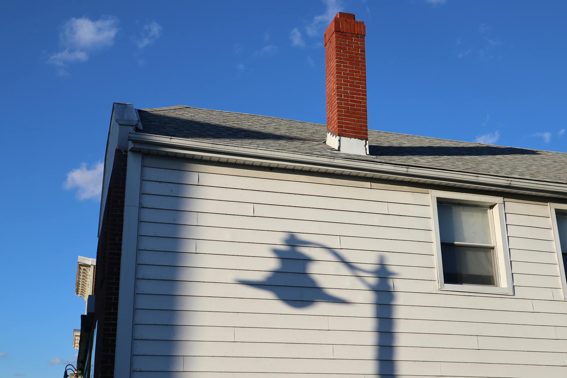 Artistic capture of a lamp post shadow on a house with a brick chimney under blue skies.